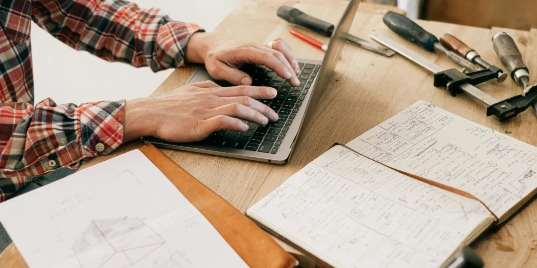 man typing on computer with construction notebook on table