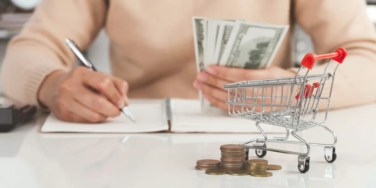 person holding money while writing in notebook with shopping cart on table