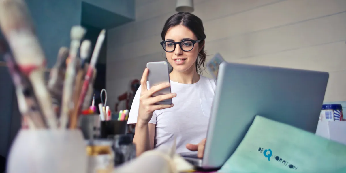 woman looking at phone with computer and art utensils on table